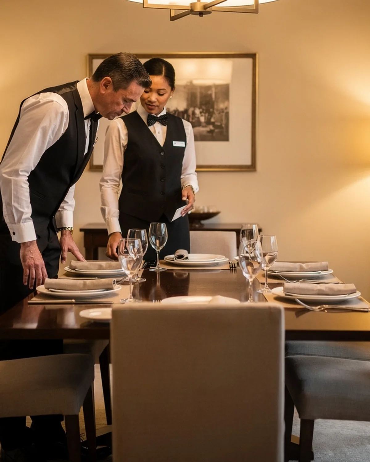 Two servers in black attire coordinating beside an elegantly set but empty dining table in a warmly lit private home dining room.